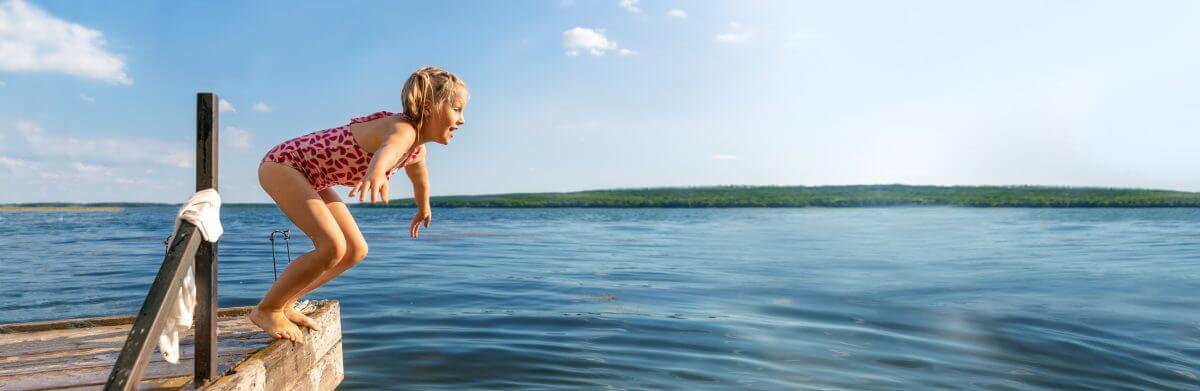 Young smiling girl with arms stretched, preparing to jump or dive off the edge of a dock into a lake. It is a sunny day with just a few clouds floating by.