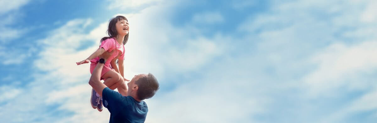 Father holding daughter in air, daughter has arms outstretched with a big small. Background is a partly cloudy sky with a majestic feel.