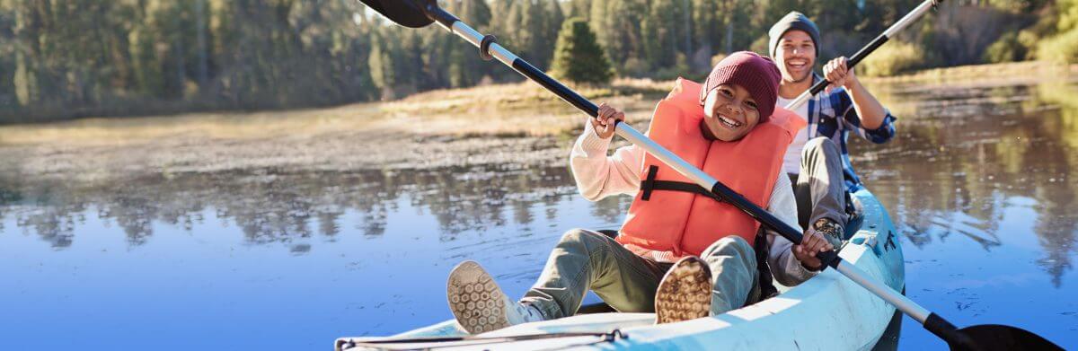 Brothers kayaking in a marshy lake with a forest background. Both are smiling while paddling and enjoying themselves.