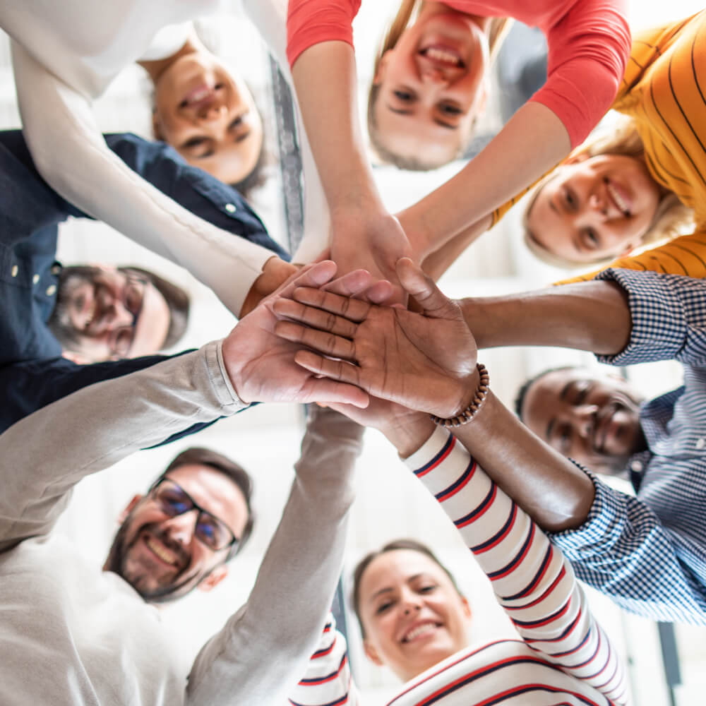 Photo looking up from the ground in the center of a team huddle. Hands are together and team is smiling looking down.