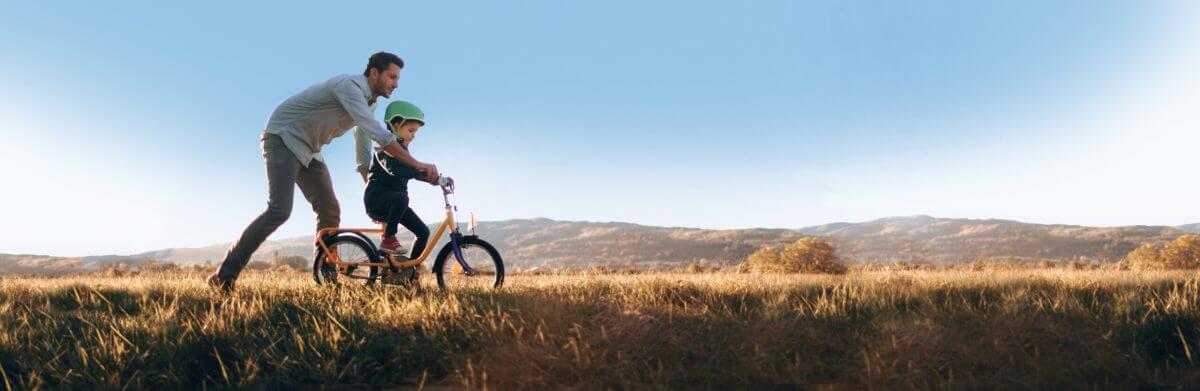Father teaching son to ride a bike. Scene is set against a hilly background with blue sky. In the foreground, a grassy plain.