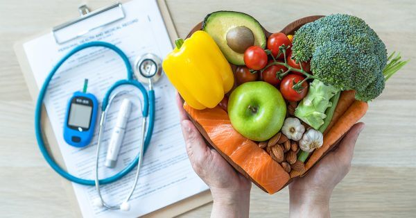 Looking down at a heart-shaped bowl of fruits, vegetables and nuts held in hands. Behind the bowl, slightly out of focus, is a medical clipboard with stethoscope and diabetes devices.