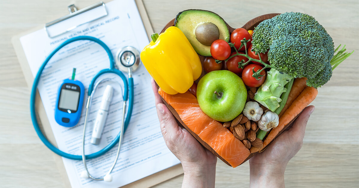 Looking down at a heart-shaped bowl of fruits, vegetables and nuts held in hands. Behind the bowl, slightly out of focus, is a medical clipboard with stethoscope and diabetes devices.